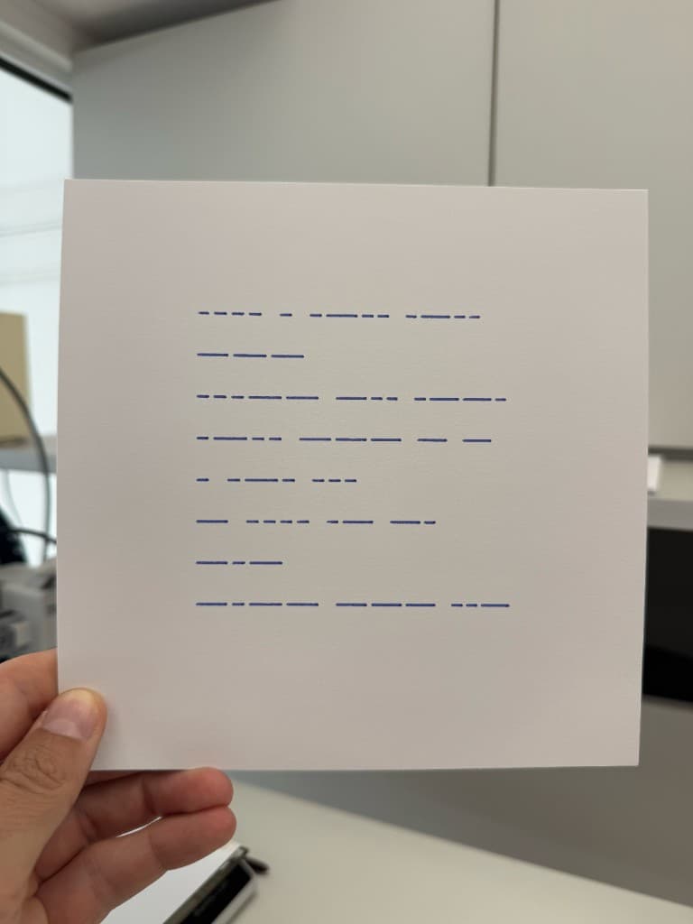 Hand holding pen-plotted Morse code on white cardstock spelling hello 3dplotters thank you in blue ink