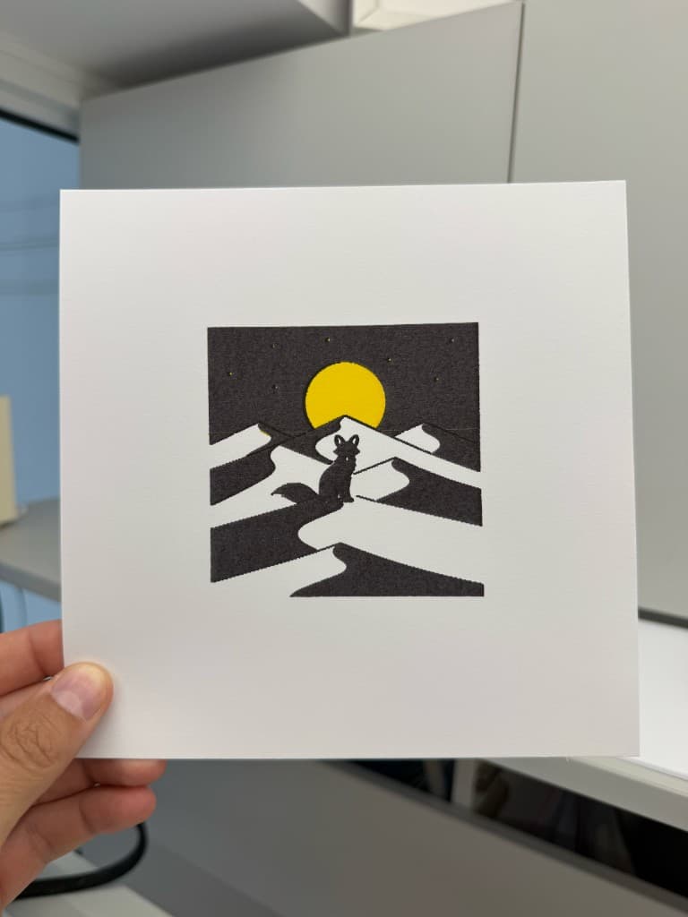 Hand holding pen-plotted GenPlot desert fox on white cardstock in black and yellow ink with moon, dunes, and stars