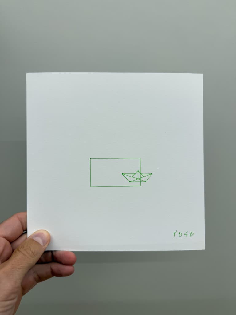 Hand holding pen-plotted green rectangle, geometric paper boat, and rose signature on white cardstock