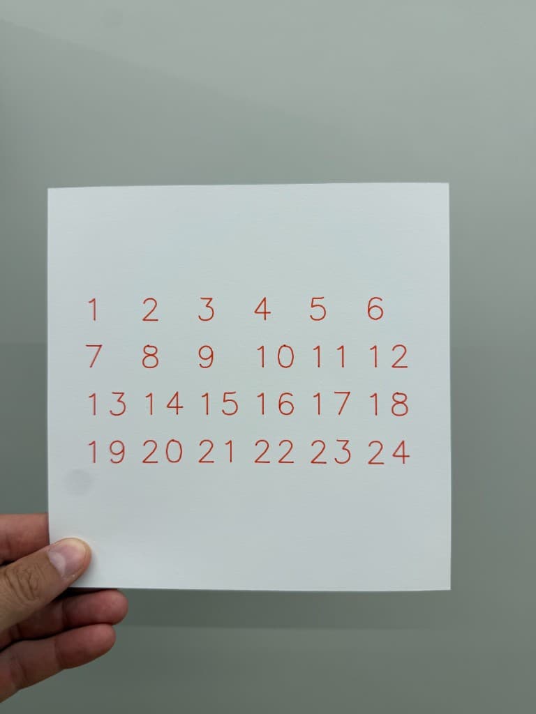 Hand holding pen-plotted numbers 1 through 24 in orange ink on white cardstock