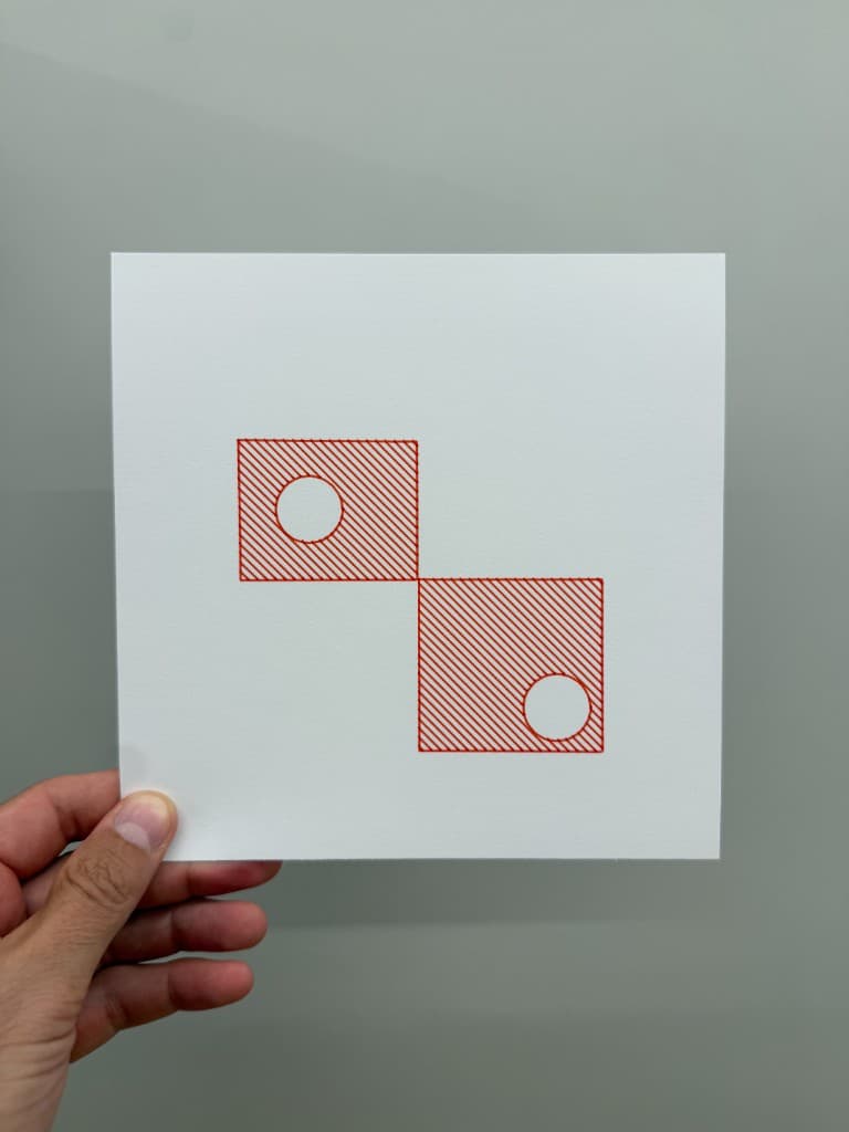Hand holding pen-plotted red diagonal hatch inside two squares with circular cutouts on white cardstock